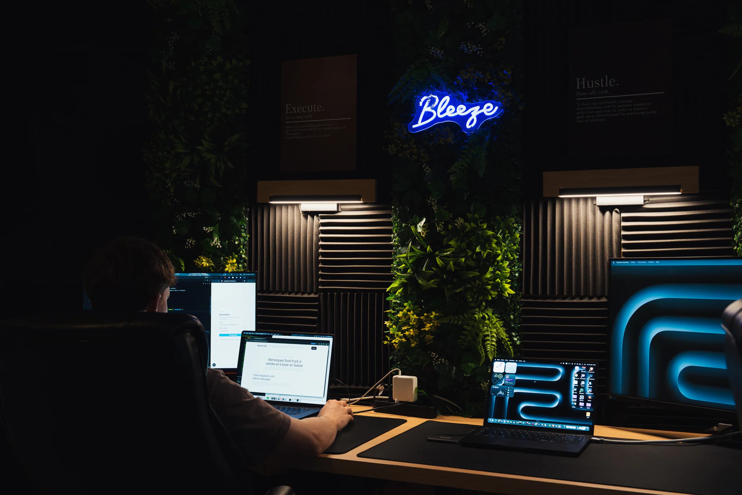 Le bureau de Bleeze : un développeur au travail devant ses écrans, sous l'enseigne lumineuse Bleeze et un mur végétal